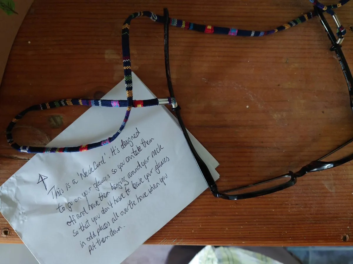 Looking down on a pair of spectacles on a shelf, with a coiled up neck cord and a note put beside them, note text in caption below image
