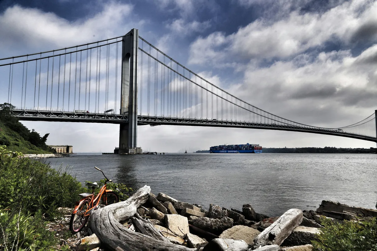 ship passing the Verrazano Narrows Bridge with an orange bicycle on the embankment in the foreground
