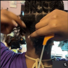A close-up, looped video of a person maintaining their microlocs. They are using the thin, metal handle of an orange rat-tail comb to precisely section the hair and guide a loc through the root area at the nape of the neck. The hair is neatly divided into a grid of small, uniform twists.