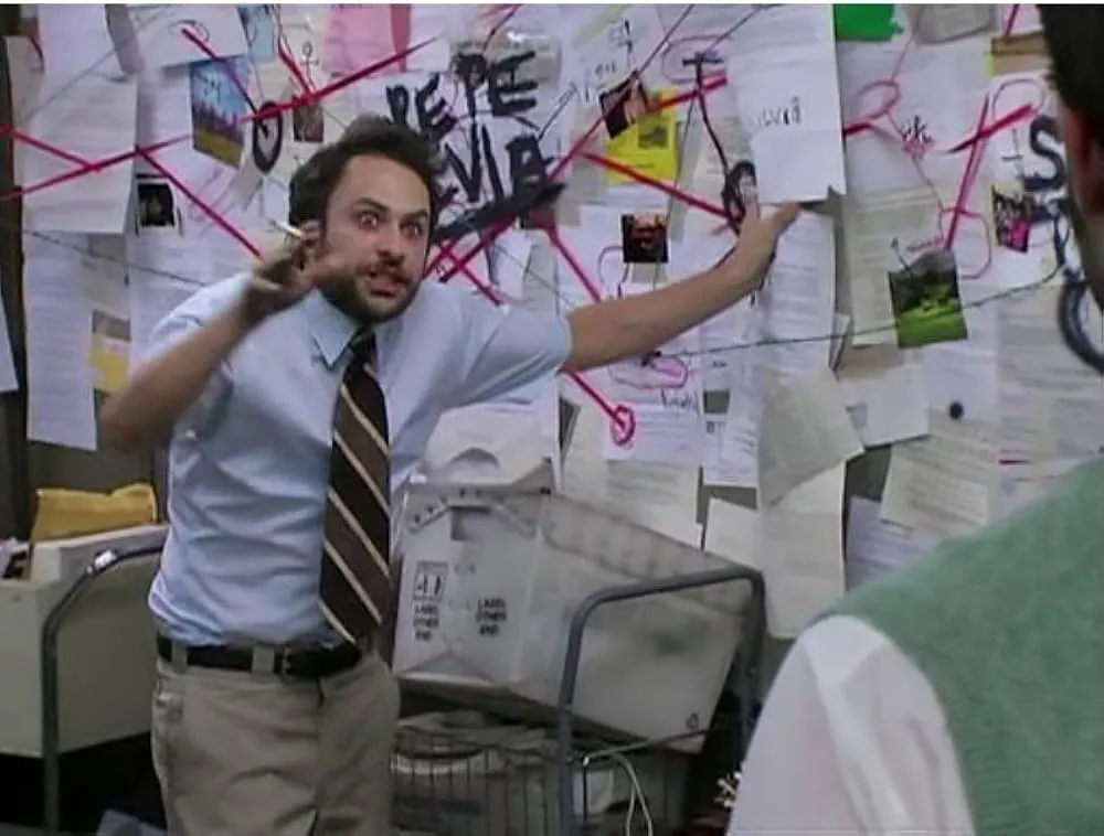 A man (Charlie Day) with disheveled hair and an intense, wide-eyed expression stands in front of a complex conspiracy board. He is wearing a short-sleeved blue button-down shirt and a striped tie, gesturing wildly with a lit cigarette in his hand. The board behind him is covered in papers, photos, and a chaotic web of red string. The words 