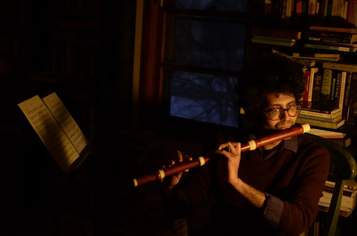 A man playing a wooden flute in a dimly lit study.