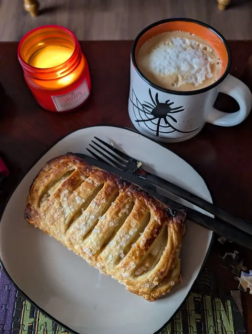 a Halloween spider mug filled with coffee, a plate holding an apple strudel, and an orange candle