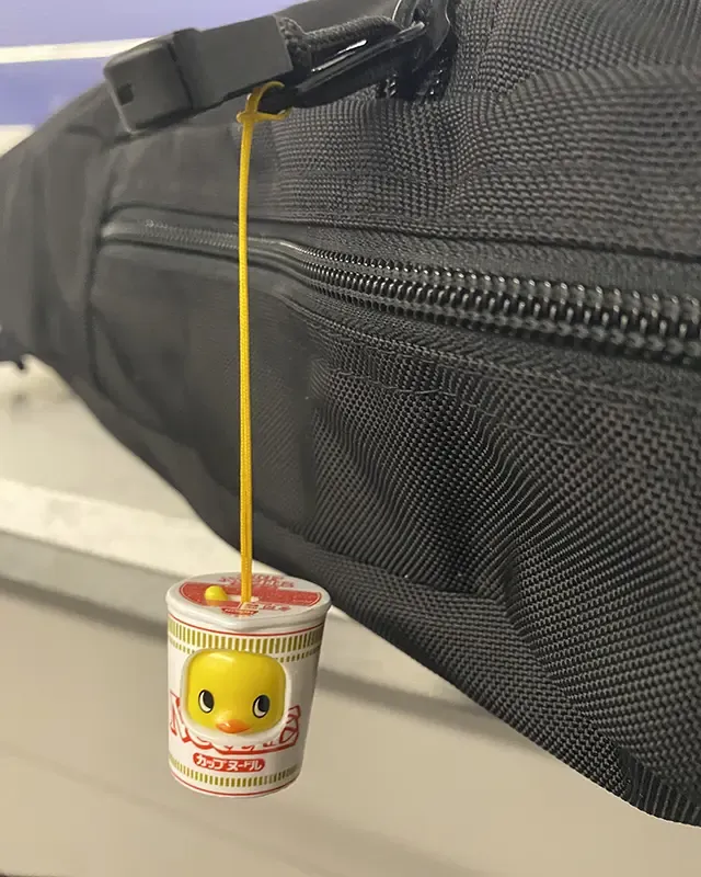 A plastic chick in a cup noodle costume hanging off the zipper of a black backpack