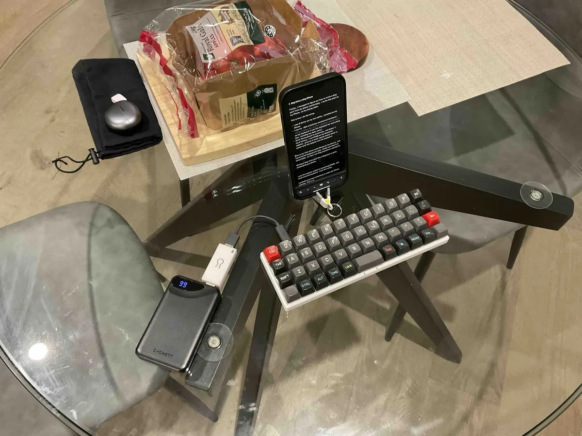 Portable blog writing setup on a glass dining table showing an iPhone on a stand displaying a text editor, connected to a 40% ortholinear mechanical keyboard via a Bluetooth adapter and power bank