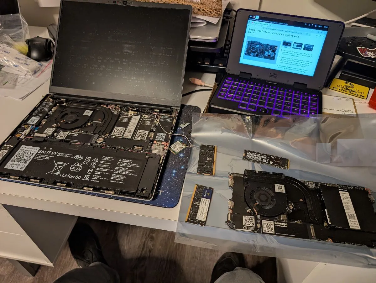 A Framework 12 laptop in the midst of a mainboard transplant on a cluttered desk. The removed Chromebook mainboard, memory, and storage modules are sitting on an antistatic bag, and the (secondhand) non-Chromebook mainboard has been installed in the open laptop shell. There’s an adorable little MNT Pocket Reform mini laptop (palmtop?) nearby displaying the Framework mainboard replacement guide.