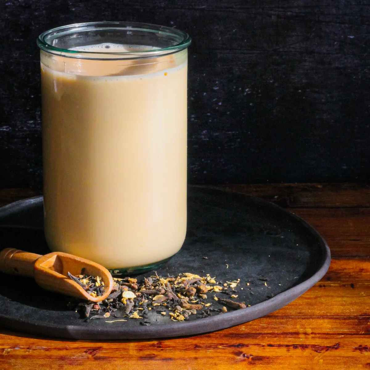 A tall glass of milky chai on a wood and iron background with a scoop of loose leaf chai.