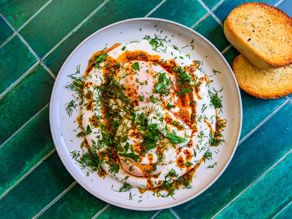 Çılbır (Turkish Eggs) on a background of green tiles with lots of green herbs on top