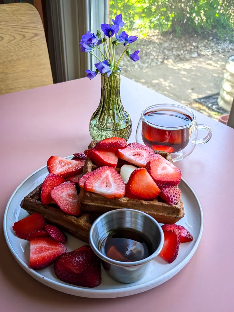 A plate of waffles with strawberries and maple syrup, a cup of black tea, and a small vase of wild violets