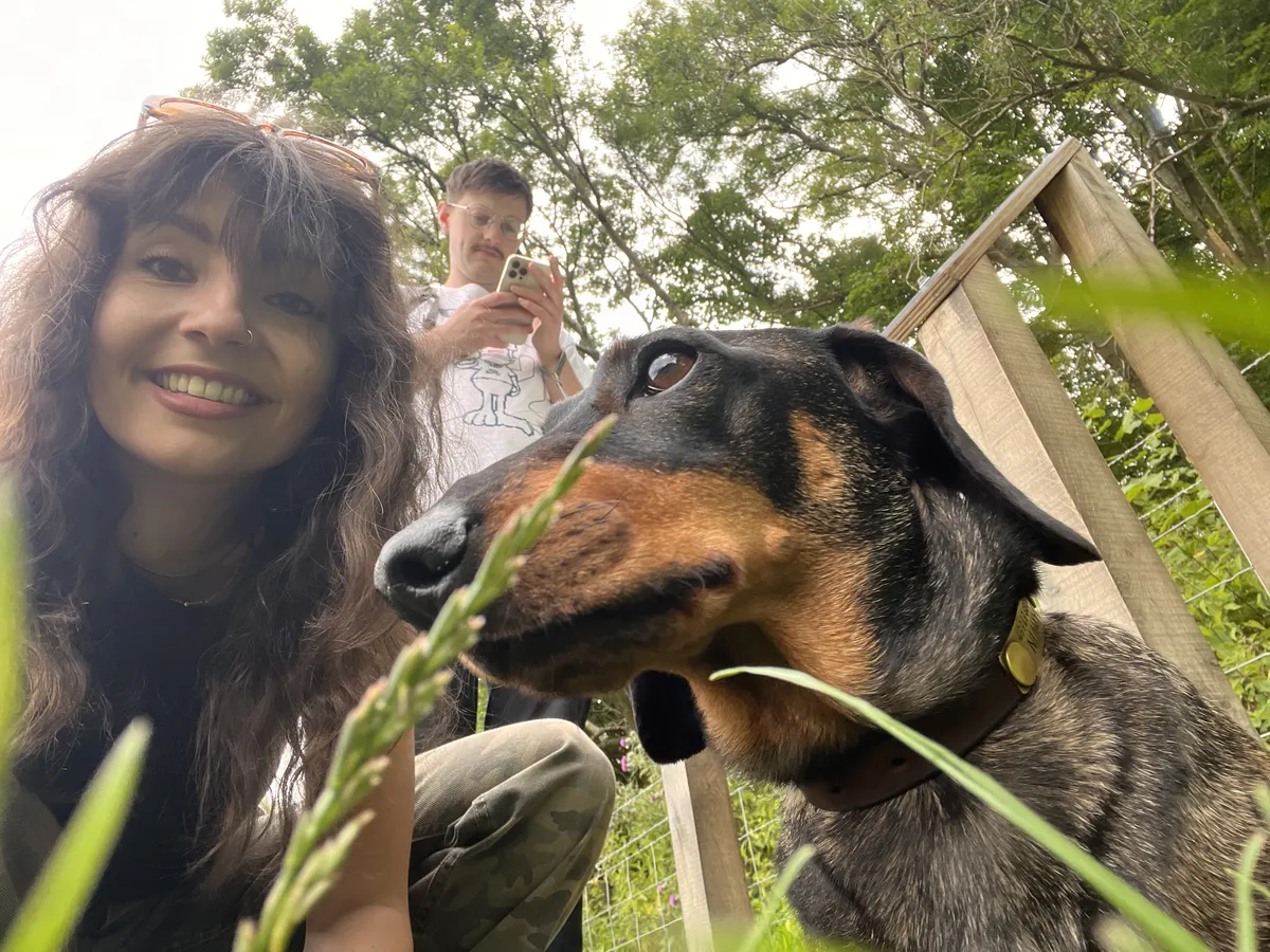 Katy, lady with brown hair smiling next to Darwin the dachshund, David stood looking at his phone in the background