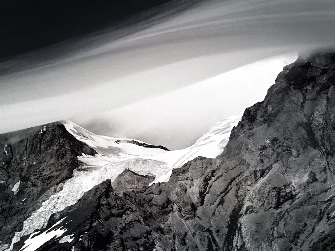 A lenticular cloud over a glacier on Mount Rainier-  a parabolic bowl with a lid of cloud. The image is high contrast, black and white. The lenticular cloud is smooth like silk and contrasts with the rough and ragged structure of the mountain and ice.
