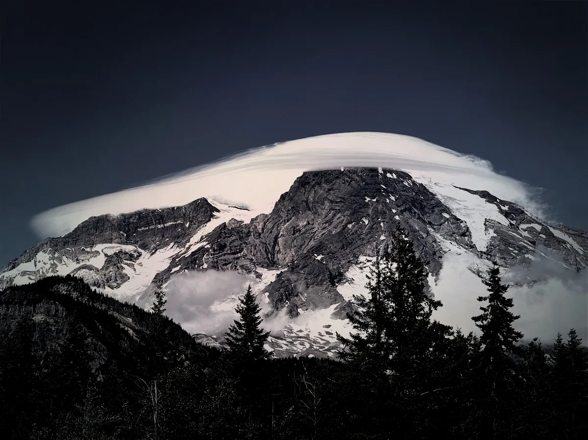 A desaturated high contrast image of Mount Rainier with a lenticular cloud that almost looks like a very shallow mushroom cap smeared around its top and around its side. There are dark, rocky textures with white glaciers and trees at the base.