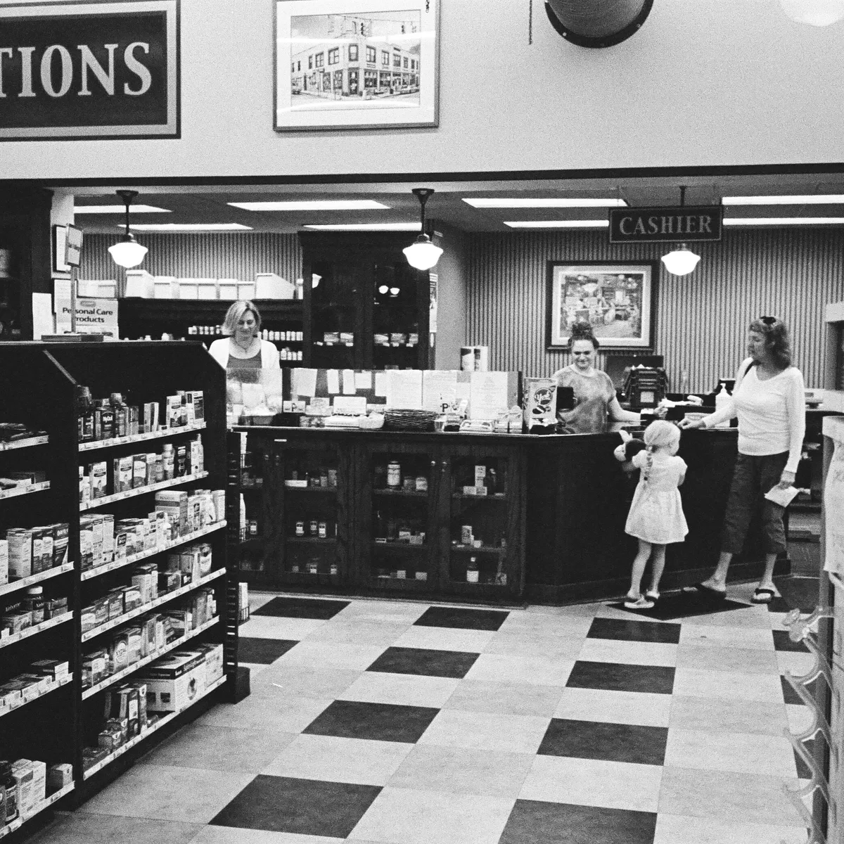 Black and white photo of a pharmacy counter in a diner where a woman and child are speaking with a cashier, with shelves of medicine in the foreground.