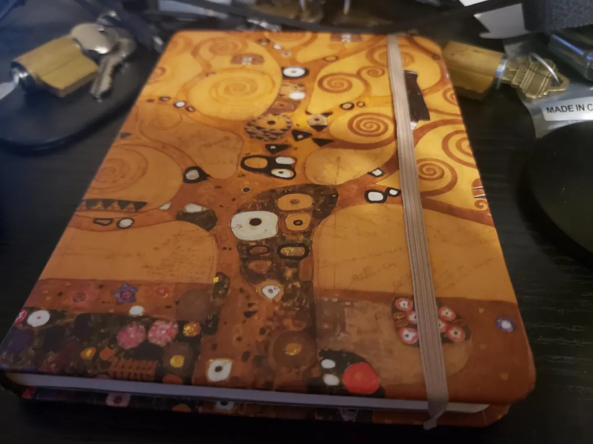 An image of a notebook on a black desk. The notebook is a kind of tannish color and has some neat-looking abstract trees on it; the desk has some scattered locks and keys lying around in the background.