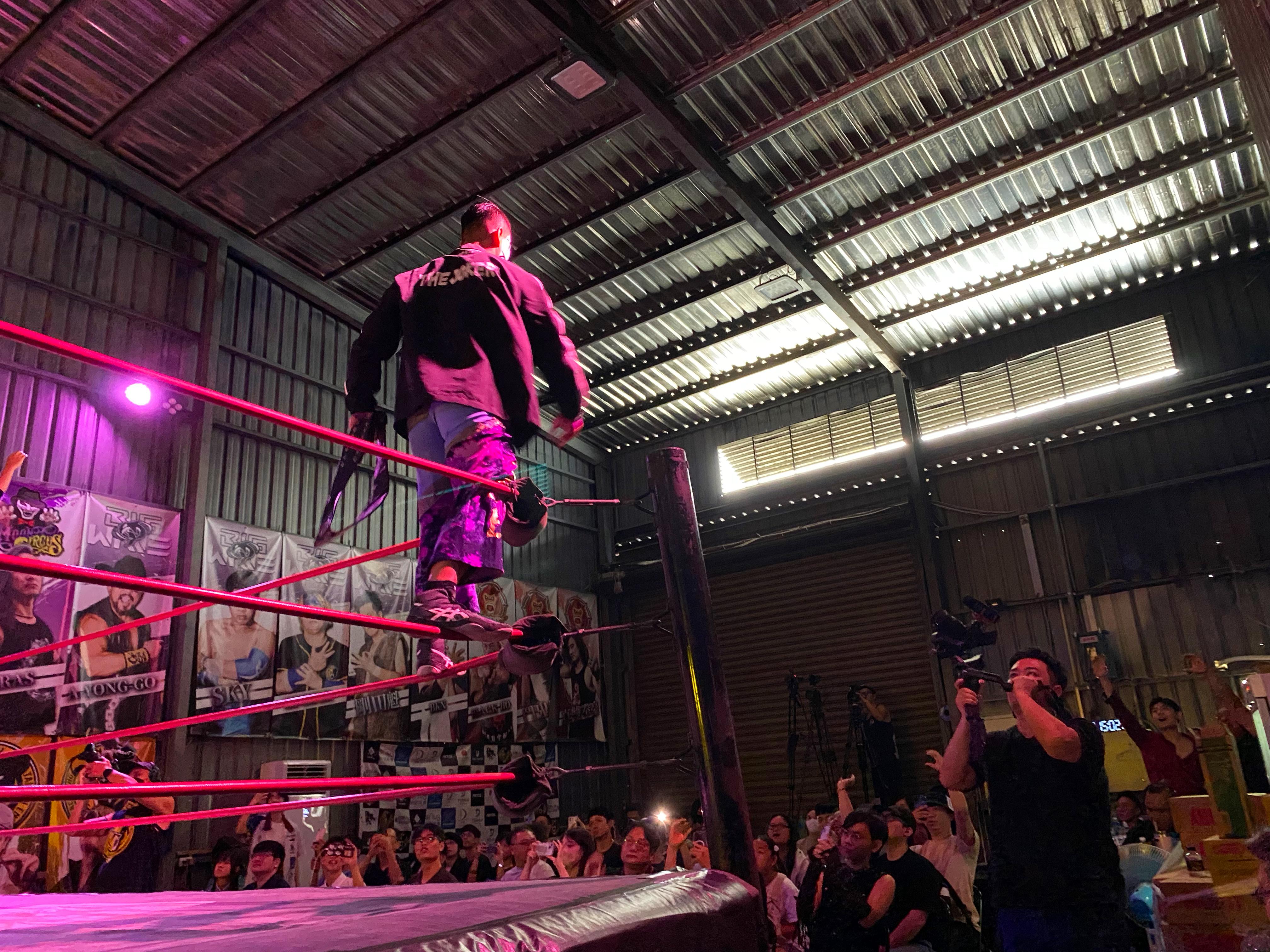 Wrestler stands on the ring post, overlooking crowd in warehouse