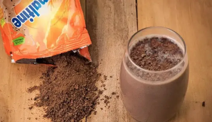 A package of Ovomaltine open and spread on a table, with a glass on the side filled with milk mixed with Ovomaltine.