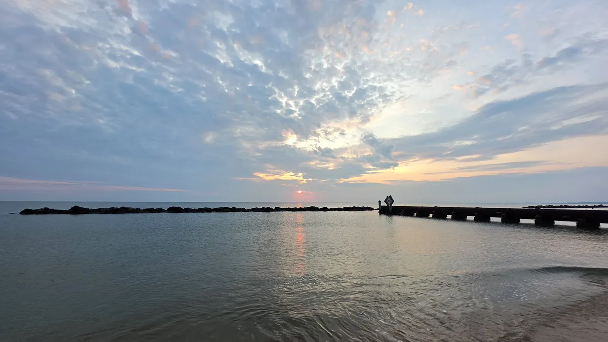 a small orange sun rises over water between grey clouded skies
