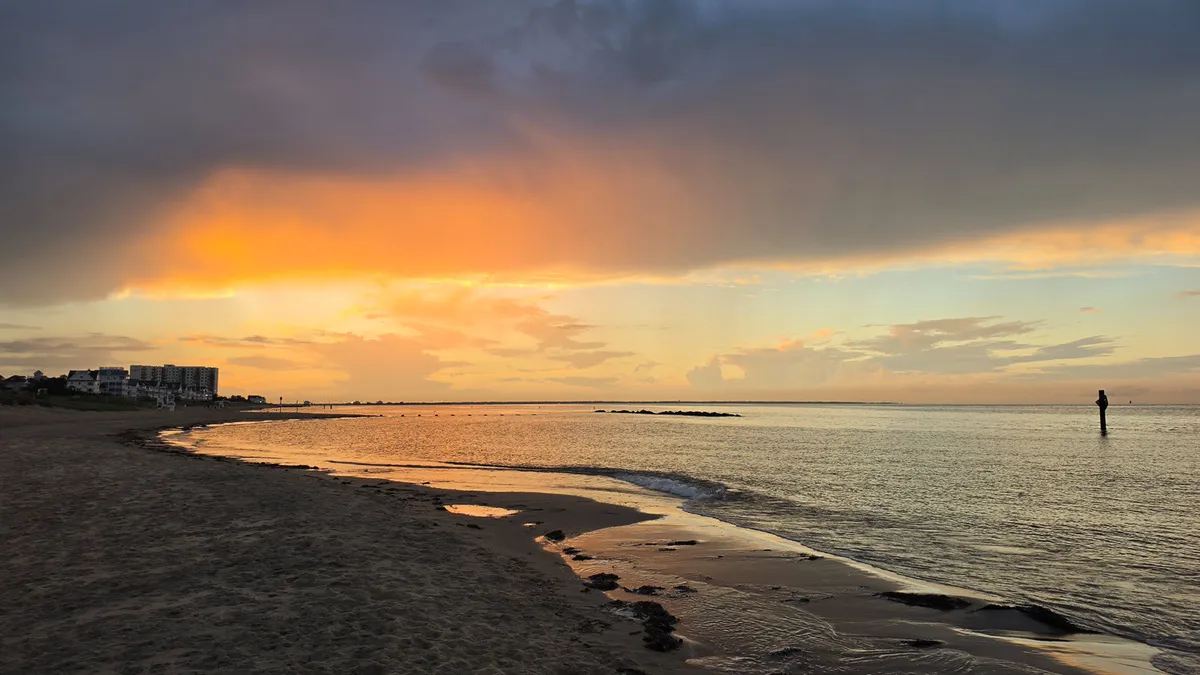orange and grey clouds obscure the sun as it sets over water and beach