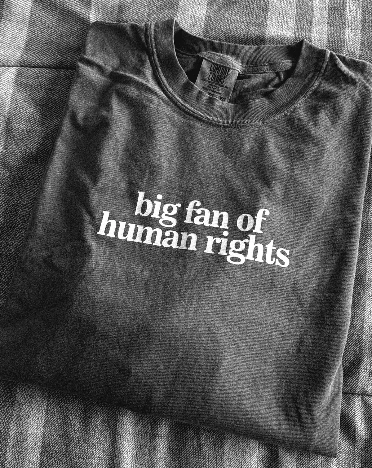 A black and white image of a t-shirt neatly folded on a bed. The chest of the shirt is screen-printed with