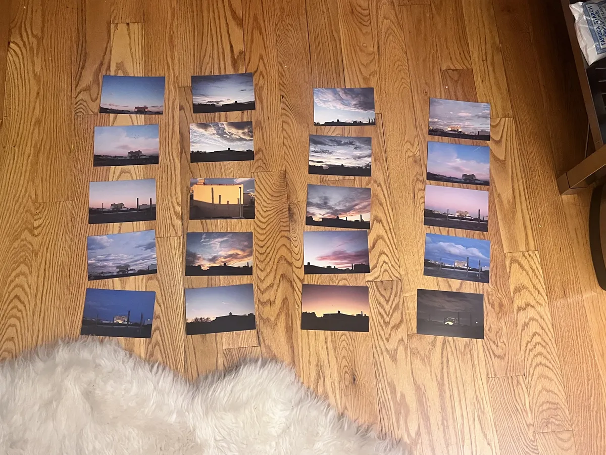 A messy grid of photos of the sunset taken from similar vantage points sits on a hardwood floor. Some of the sunsets are bombastic, others just muted gradients. The photos range in brightness and weather.