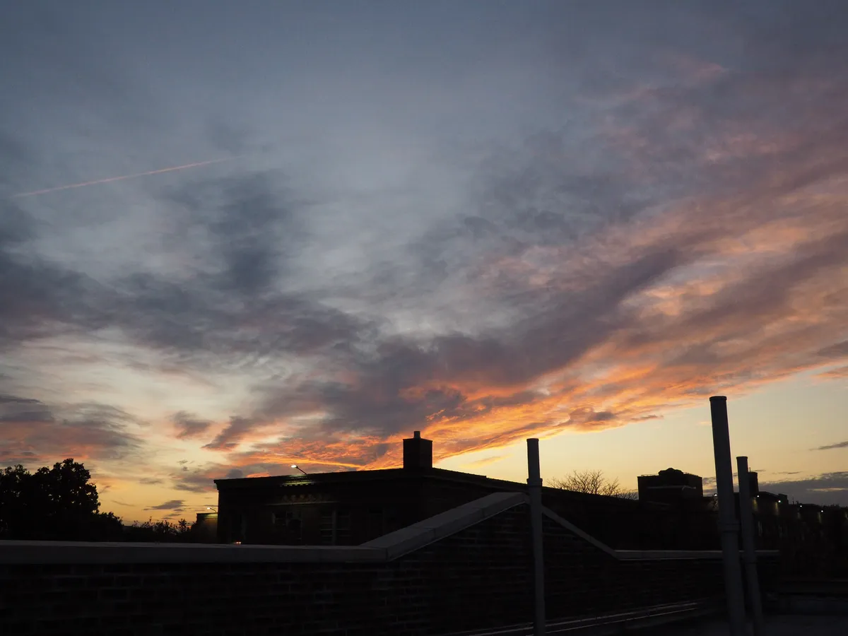Several chimneys silhouetted against a splash of dark gray clouds tinged fiery orange by the sunset. The sky is a gradient from goldenrod to light gray in the background. A plane contrail is visible, faintly glowing red against the clouds. 