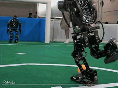 A humanoid robot kicks a soccer ball towards another humanoid robot who is standing in front of a goal. The robot goalie falls over entirely to prevent the ball from going in the goal.
