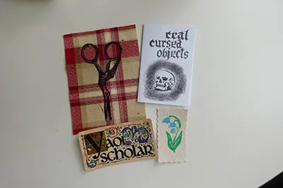 A plaid fabric swatch with an ink print of scissors, A mini zine showing skull, A rectangle sticker that says 'Yaoi Scholar', A small patch with scalloped edges with a flower printed.