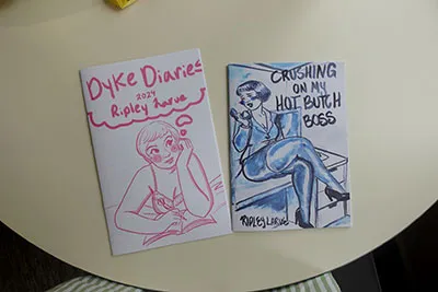 Two booklet zines. The left zine is a woman depicted in pink ink and the right zine is a woman depicted in blue ink.