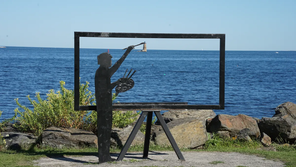 A metal frame depicting an ocean painting surrounds open water, with a metal sculpture of a painter appearing to paint Whaleback Lighthouse into their artwork.