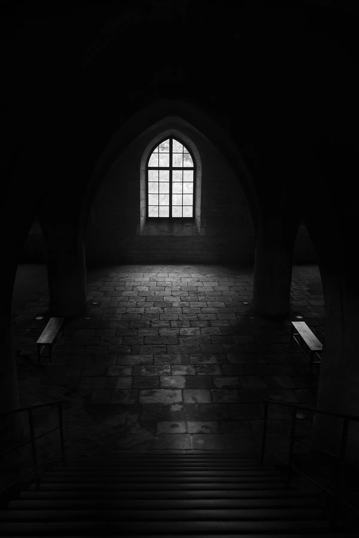 Dark, moody black-and-white photo of a vaulted stone crypt or chapter room, lit by a single arched window casting light onto the paved floor, with wooden benches beneath the pillars on either side.