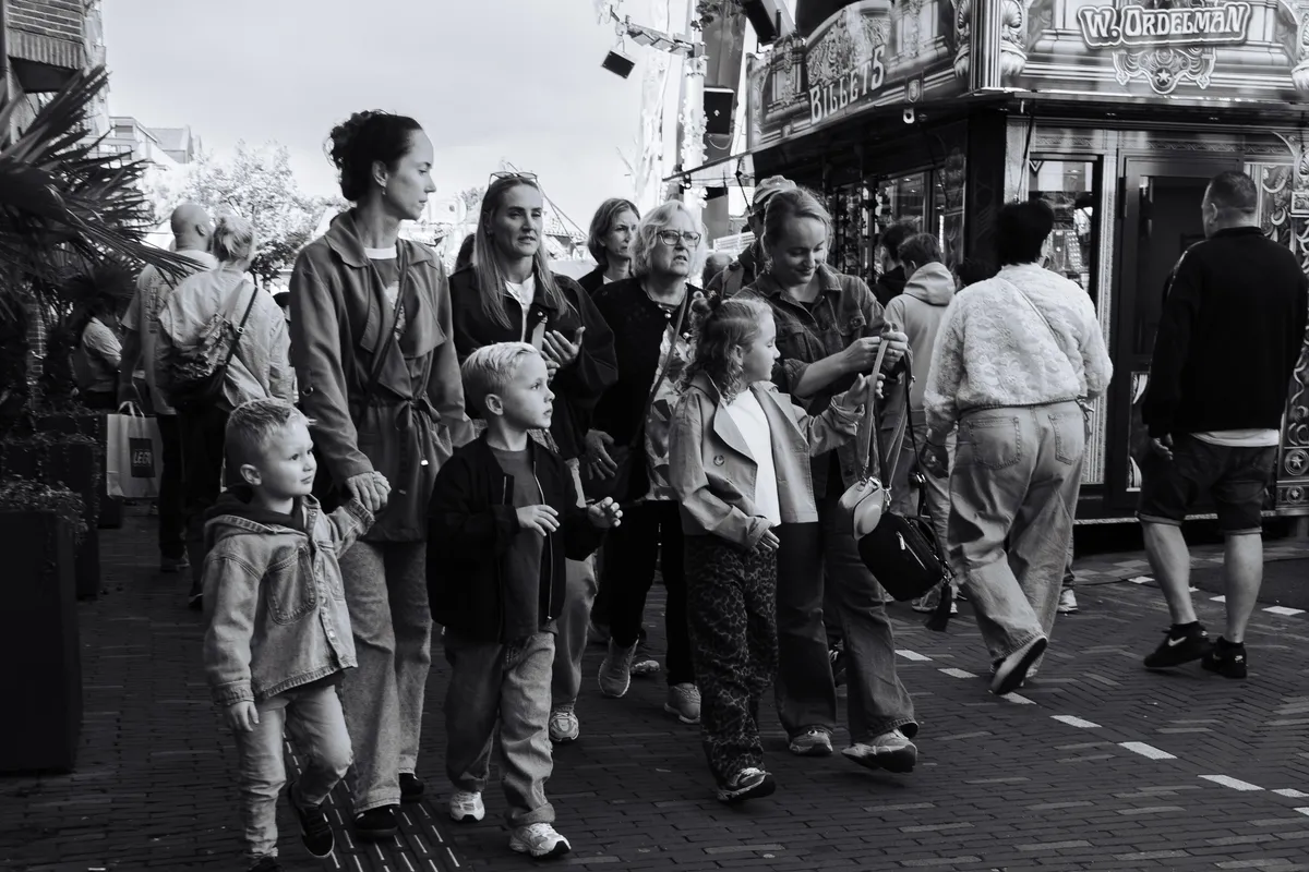 Black-and-white street photo at a fair: two young boys look off to the left, a girl holds a small handbag while a woman fastens its strap, and several women walk alongside them past a fairground stall marked Billets.