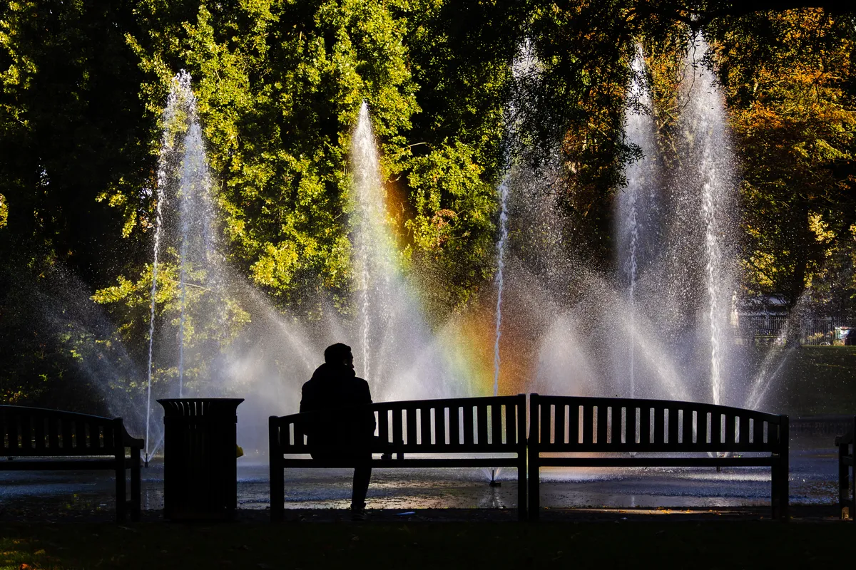 A person sits on a park bench, seen from behind in silhouette, facing a row of tall fountains. Sunlight catches the spray and forms a faint rainbow, with green and orange autumn trees behind.