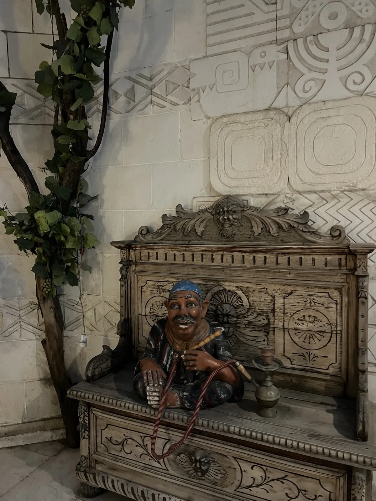 Small statue of a man with a moustache holding a shisha pipe, seated on a bench