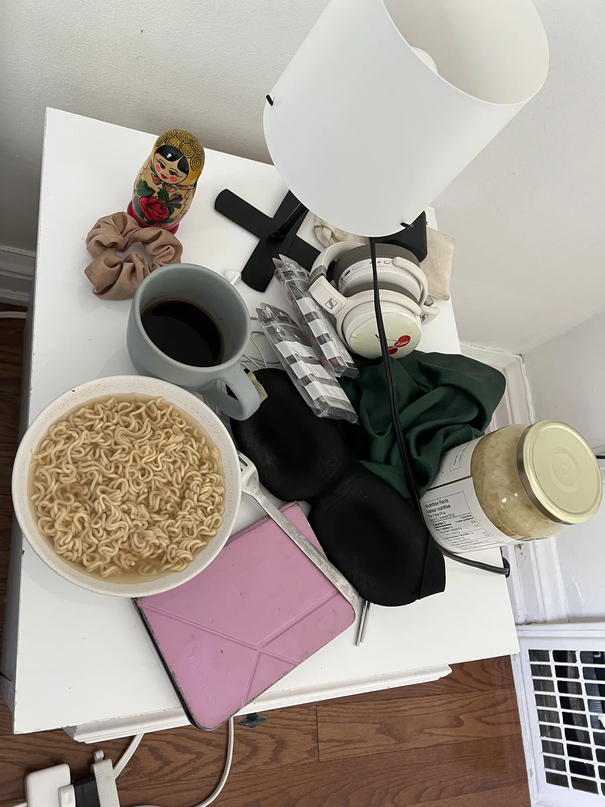 Messy bedside table littered with items that provide me with sustenance: Instant noodles, black coffee, Oratene pills, eye mask, kobo, headphones, sauerkraut