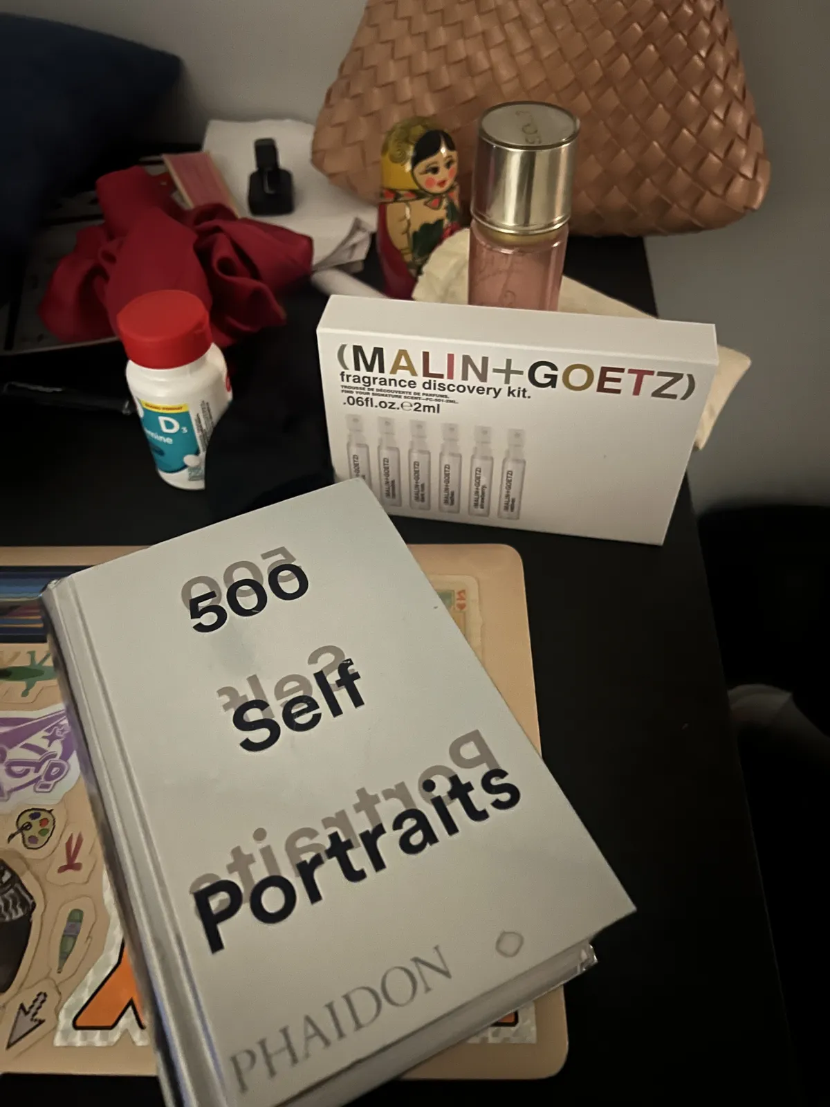 Part of my desk showing a library book, laptop, pink purse, perfume, russian doll, scrunchie, vitamins